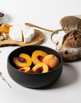 A black stoneware serving bowl filled with assorted fruits, placed on a neutral background with additional food items arranged around it.